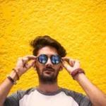 asual portrait of a man adjusting sunglasses in front of a yellow wall